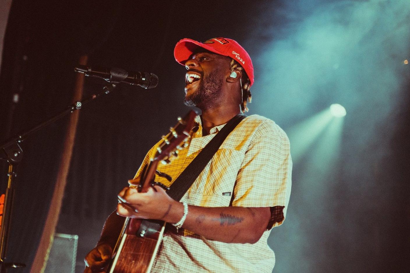 Myles close up from the side on stage, singing into a mic and playing his guitar. He wears a red baseball cap and short sleeved checked cream shirt. Dry ice smokes from above with a light shining on him.