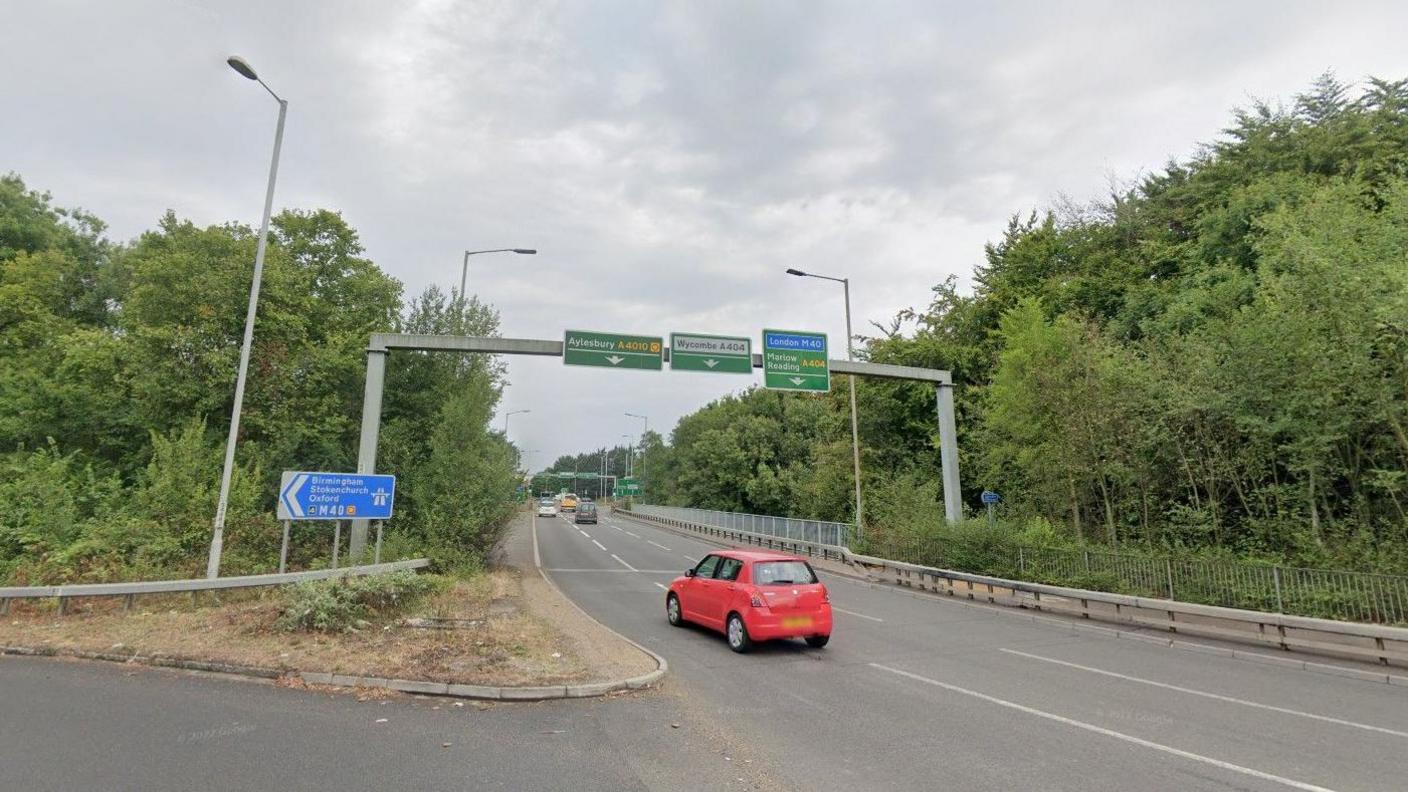 A red car is in the foreground going round the Handy Cross roundabout near the south bound entry road to the M40. The road is tree-lined with signposts overhead.