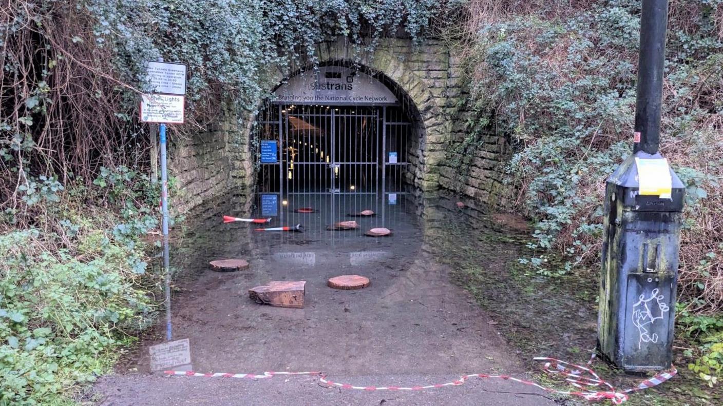 The closed metal gate at the entrance to the Devonshire Tunnel. There is red and white tape laying in the water on the ground, which had been used to block off the tunnel. There are two orange cones lying on their side in the water, and you can see dim orange lights reflecting on the water that has flooded the tunnel. 