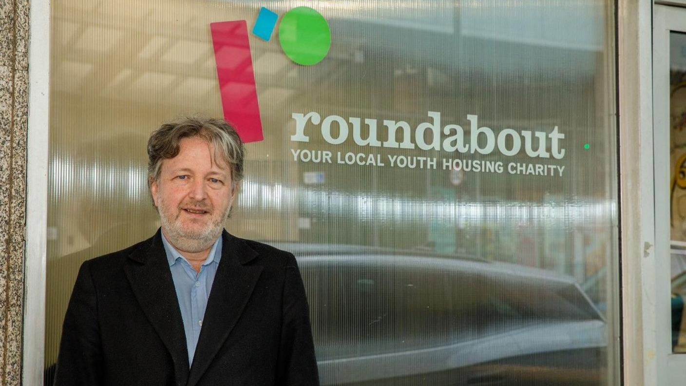 Ben Keegan stands smiling in front of a glass window with the charity logo that reads "roundabout, your local housing charity".