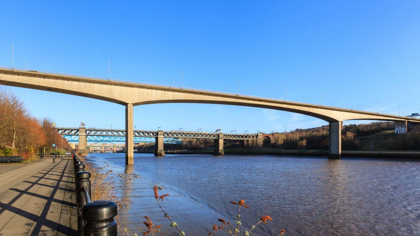 The Redheugh Bridge spannign the River Tyne. The concrete bridge is made of three shallow arches. Two piers supporting the bridge stand in the river. The picture has been taken from a footpath on the Newcastle side of the river.