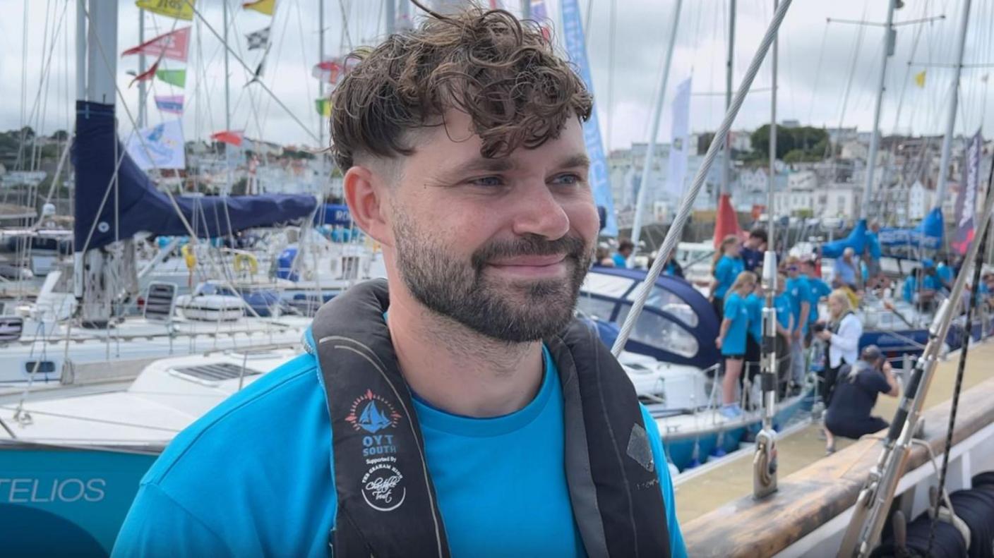 Joshua Caudary - man with curly brown hair shaved sides, brown beard smiling with blue eyes wearing a blue shirt and a small black life vest, background blurred of loads of boats on a grey sky day