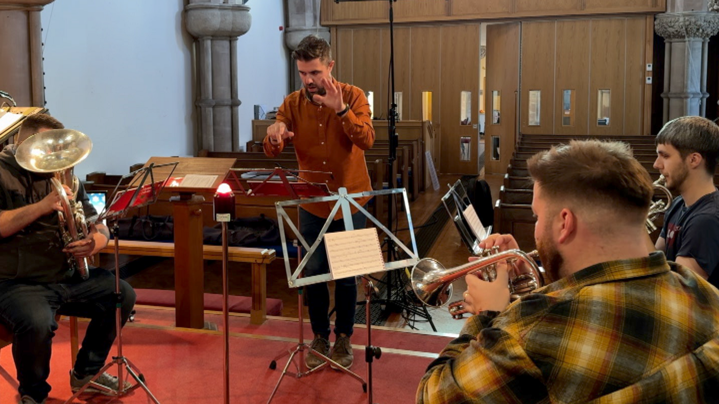 Three seated musicians playing brass instruments in a church.  They are being conducted by a man standing in the centre of the group.