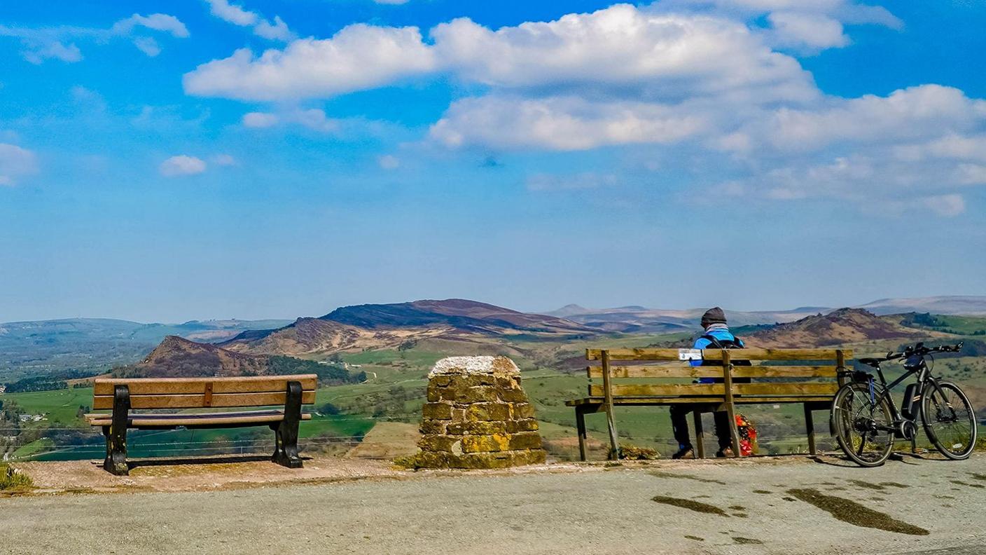 A person sits facing away on the right hand of two benches. They wear a woollen hat and a blue top with the straps of a rucksack visible on their shoulders. A circular marker made of stones sits between the benches and a bike rests against the back of the bench the person is sitting on. In front of the benches, fields and hills roll away to the horizon.