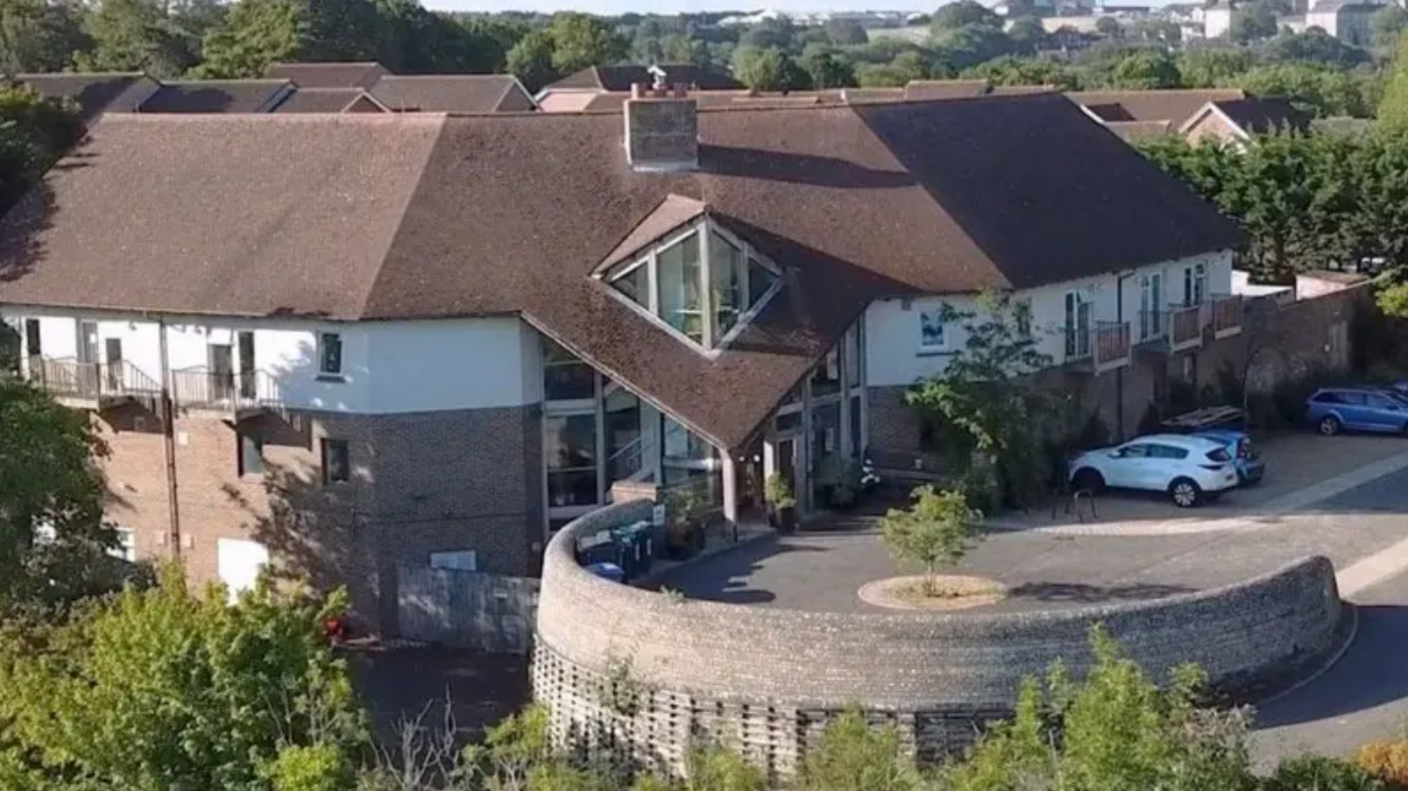 An aerial view shows The Sussex Beacon on Bevendean Road in Brighton. Leafy trees line the building's perimeter with multiple cars parked outside the building's reception.
