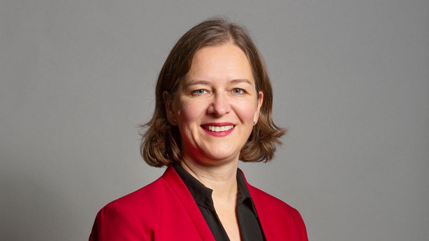 Parliamentary photo of Fleur Anderson wearing a red jacket and black shirt stood in front of a grey background