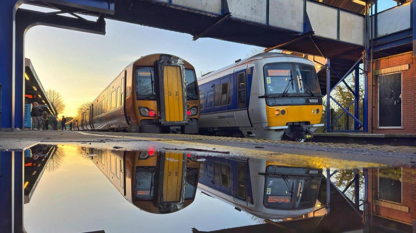 Two trains next to a railway station platform which has a puddle on it in which they are reflected.