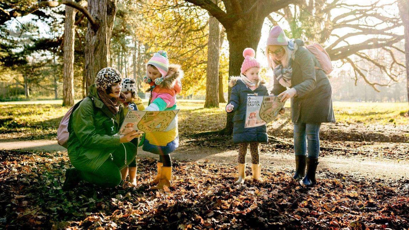 Two women and three young children wrapped up in winter coats and hats and gloves on a festive trail in a park reading a map on a sunny day. 