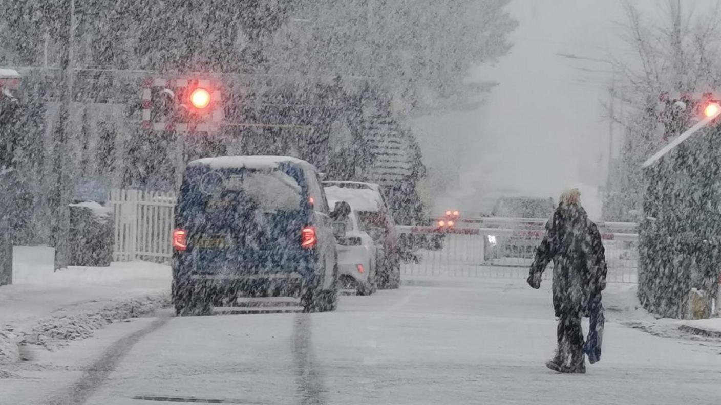 Snow falls in the village at a railway crossing. The barriers are down and warning red lights are on. There is traffic parked on either side of the barrier.