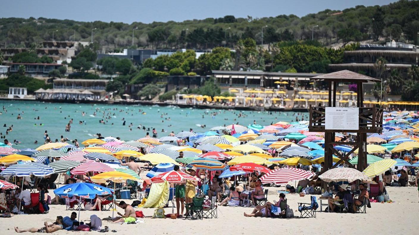 A summer beach scene with hundreds of people under sun shades and a blue sea