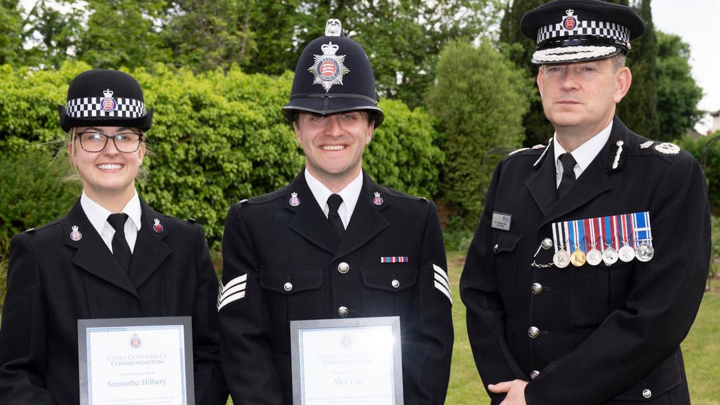 PC Sam Hilbery and Sgt Alex Farr with Essex Chief Constable Ben-Julian Harrington, all in police uniform, facing the camera, they all have police hats on. Sam and Alex are holding certificates in a frame.