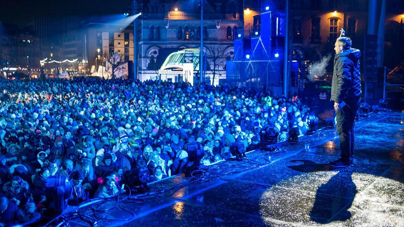 Steven Frayne on stage wearing a puffer coat and jeans speaking to an audience of thousands in Bradford's City Park.