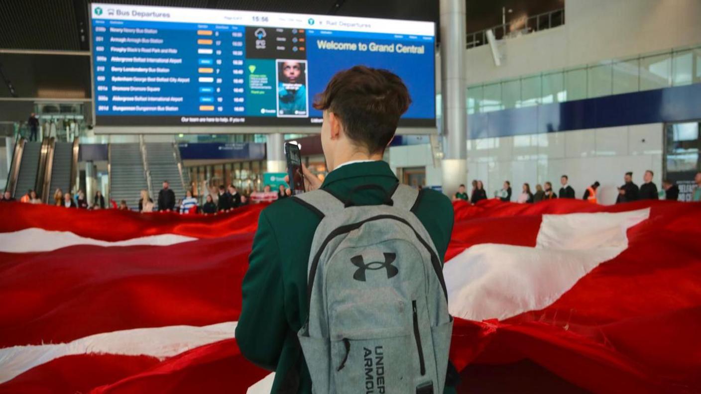 A boy in a green blazer with his back to the camera with a rucksack on in front of a red and white flag