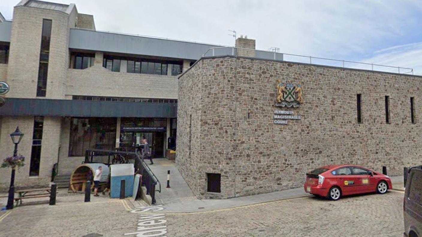 The front entrance to Plymouth Magistrates' Court. It is a stone building on a cobbled road, with a ramp built leading to the entrance. The logo can be seen on the front of the building.