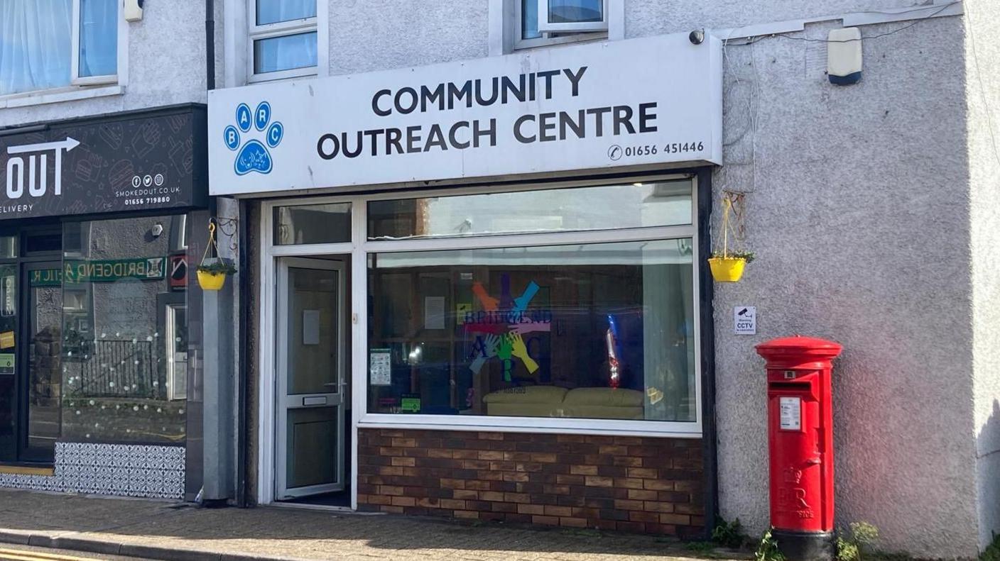 A building on a street with a big glass window at the front and a white sign which reads Community Outreach Centre. There is a red post box next to the window in front of a grey wall and a yellow hanging basket