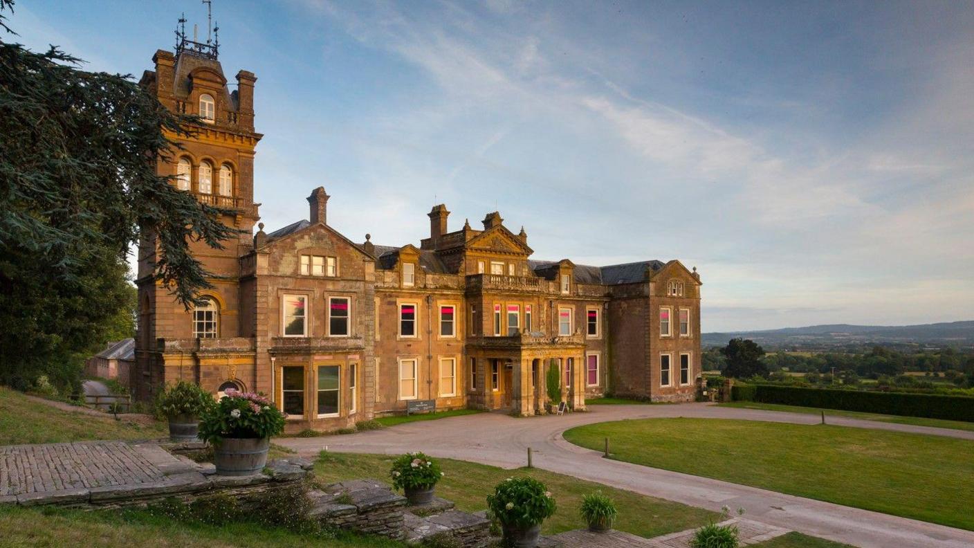 The front aspect of Hestercombe House, a grand, beige-gold stoned building that has a tower to the left. In front of the house is a large driveway with an oval of grassy lawn. Fields and trees extend into the distance.