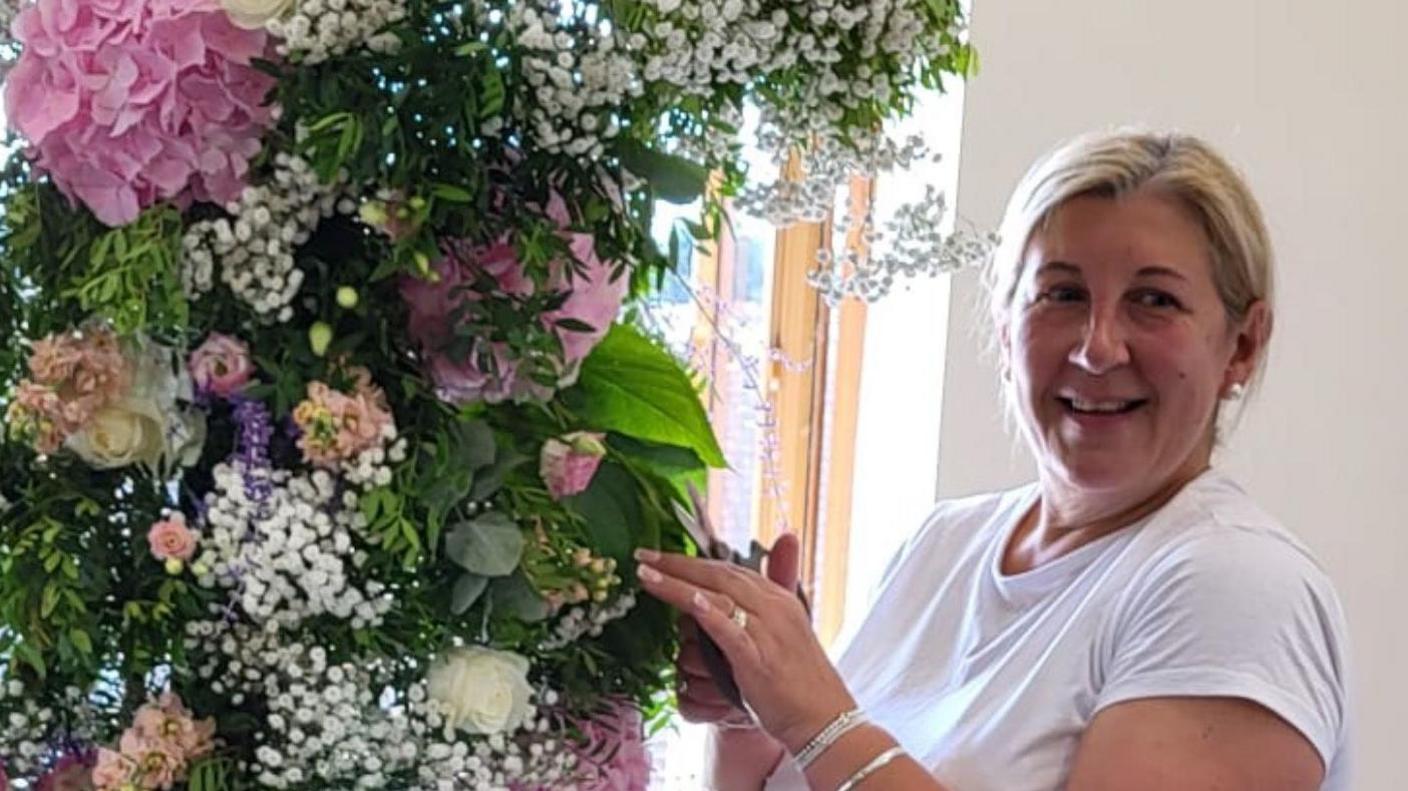 Leah Francombe, 58, has blonde hair, tied back and parted in the middle of her forehead. She has pearl earrings and some silver bracelets around her left wrist. Both her hands are touching the leaves on a large bouquet of flowers. Behind her is a white wall with a window frame visible too.