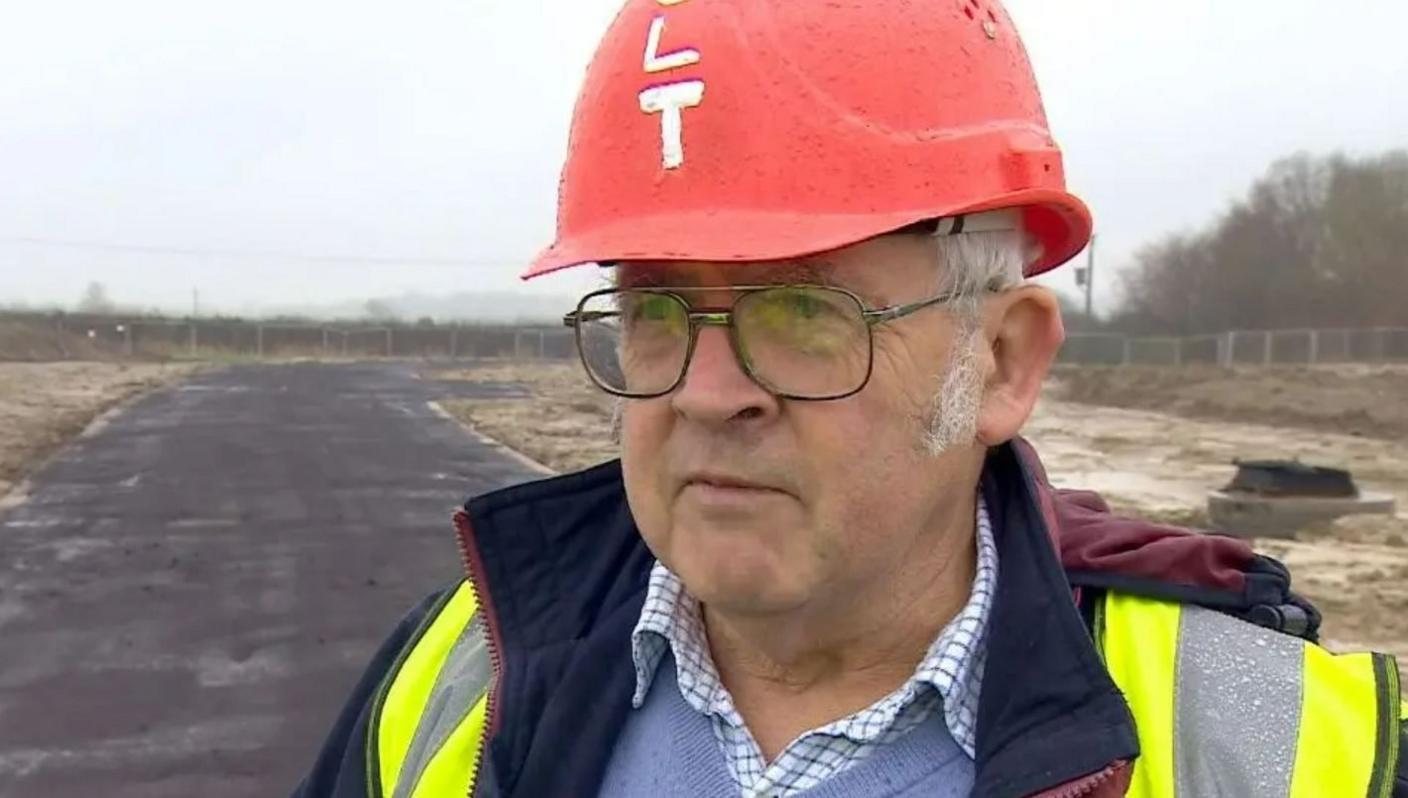 Tom wearing red heavy hat, glasses and high vis jacket on building site with long road of tarmac.