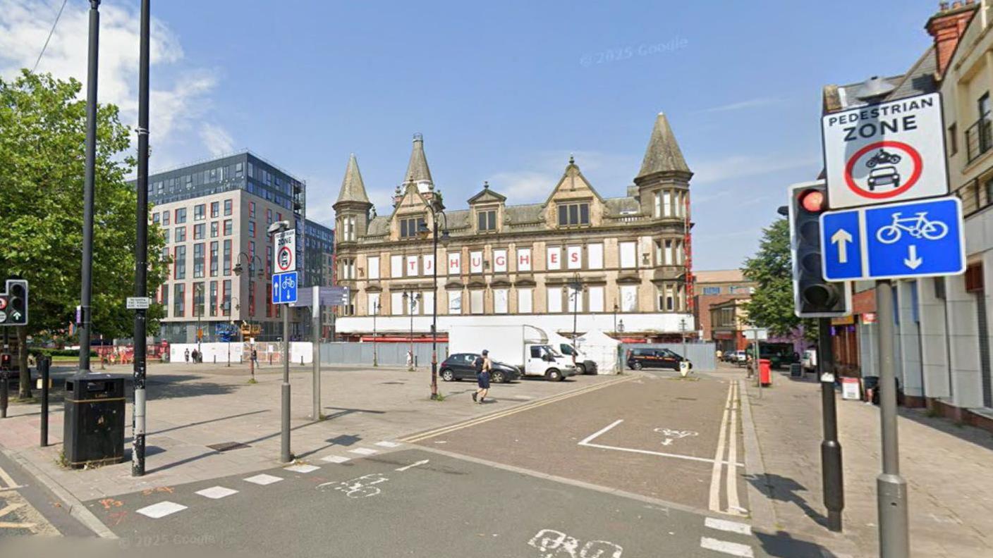 A large paved area with trees on one side and a boarded up yellow brick building which has a sign in red lettering which reads TJ Hughes. A white road sign with black writing which reads pedestrian zone and a blue road sign showing a cycle lane and an image of a bicycle and arrows are next to a set of traffic lights.