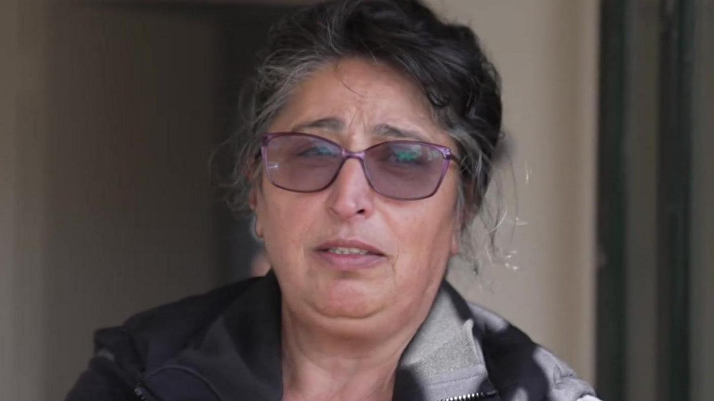 A woman with greying dark hair pulled back, wearing purple glasses and a black jacket, looks directly at the camera. The background is out of focus, showing a plain wall and doorway.