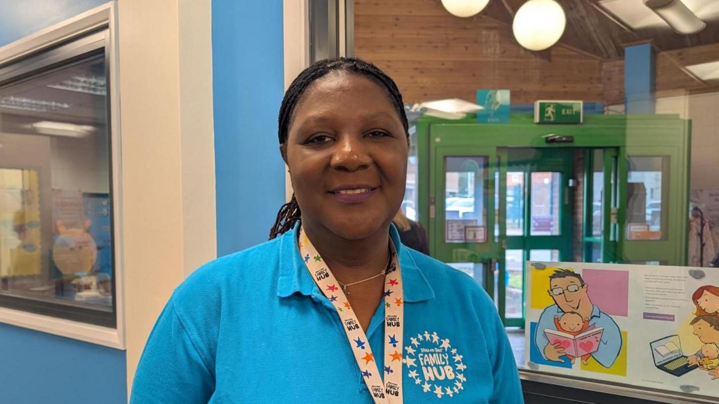 A woman wearing a bright blue polo shirt and colourful lanyard, wither her hair tied back, smiles at the camera. She is standing in a foyer area, with colourful pictures on a display and a green fire exit door behind her.