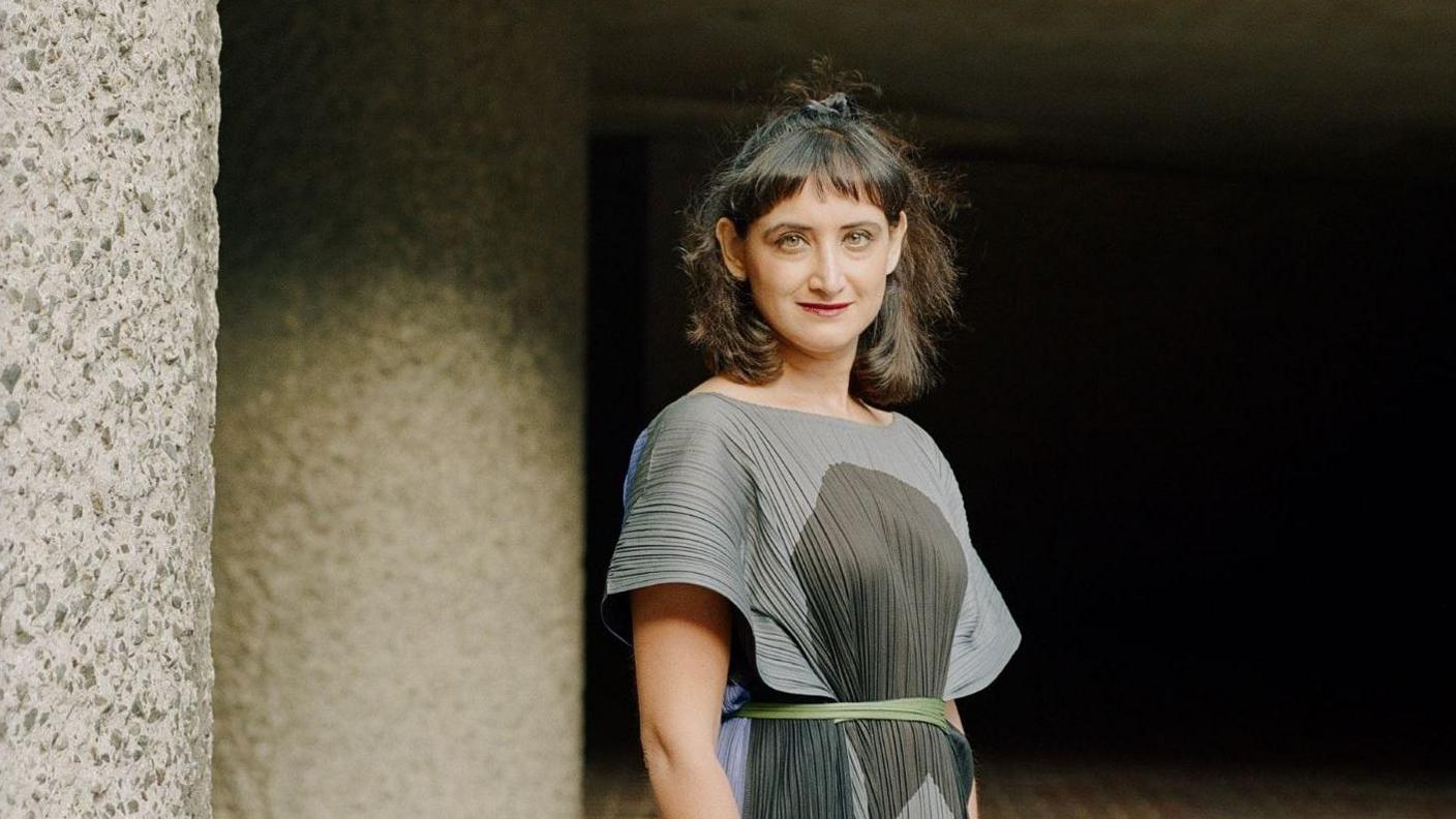 A woman wearing a grey and black dress stands outside. She has black hair tied back and a fringe. There are granite walls in the background.