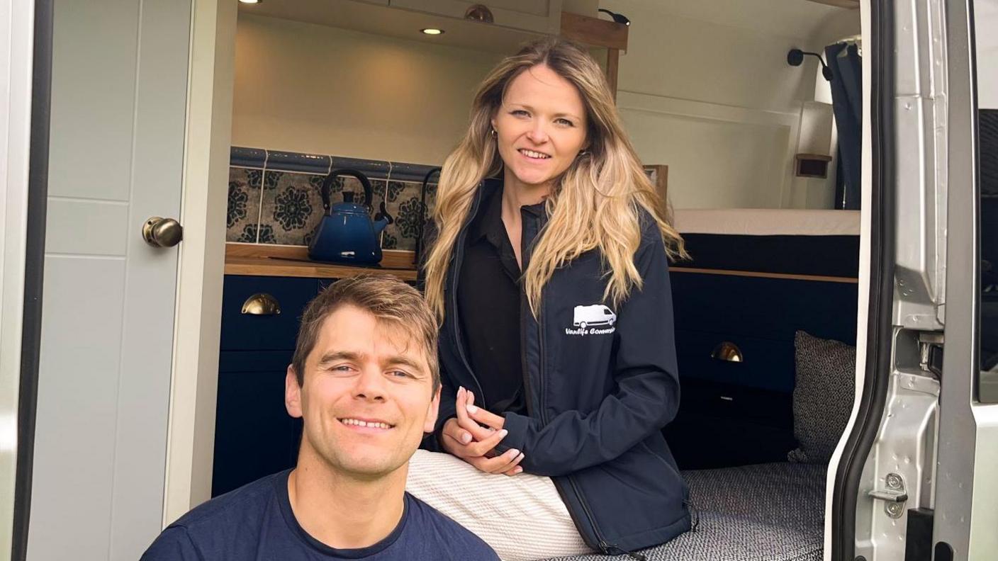 Mr and Mrs Arnold sitting in a converted van and smiling. The van has a worktop, bed and kettle pictured. The couple are wearing navy company clothing.