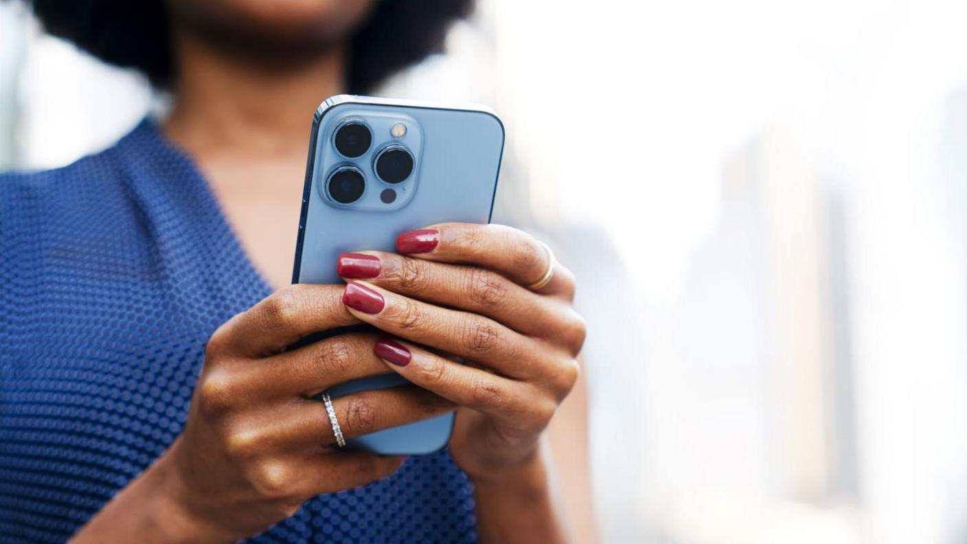 A woman is holding a mobile phone. She has her nails painted burgundy. She's wearing a ring and has a blue top on. 