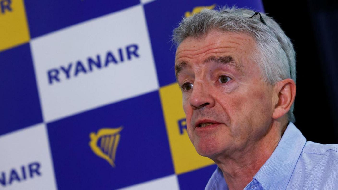 To the left of the photo is Michael O'Leary, pictured from the shoulders up. He is wearing a blue shirt and has grey hair. In the background is a poster with the word Ryanair and the logo on it. 