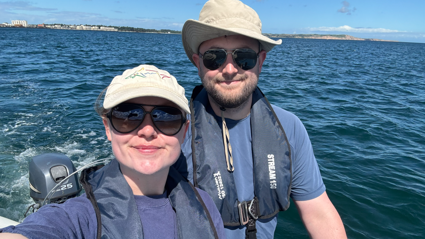 Gemma Scotts (left) and Tom Morgan are on a boat wearing small blue life jackets around their necks, wearing sunglasses and beige hats. A vast sea is behind them with a distant coastline on sunny day.
