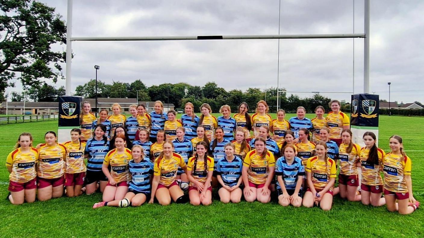 Two girls rugby teams pose for a team photo under the posts. They are wearing a mix of yellow and blue jerseys and are standing on a grass pitch. 