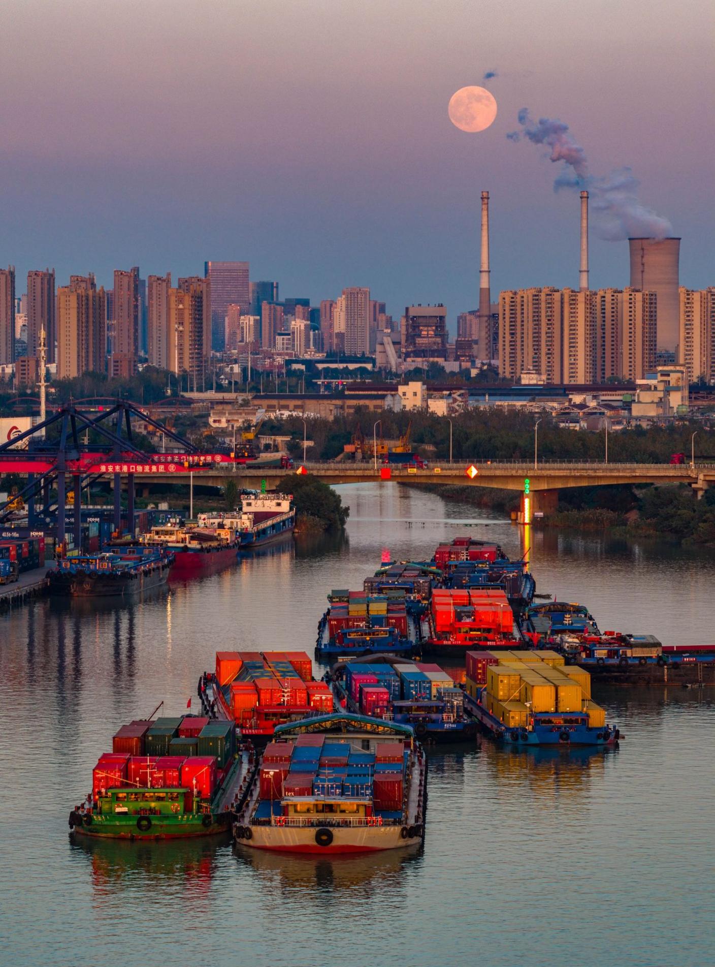 Freight ships are seen moving down the canal in front of a bridge and the city skiyline, with power plant chimneys smoking next to the Moon rising over the city, in Huai'an city on Wednesday.