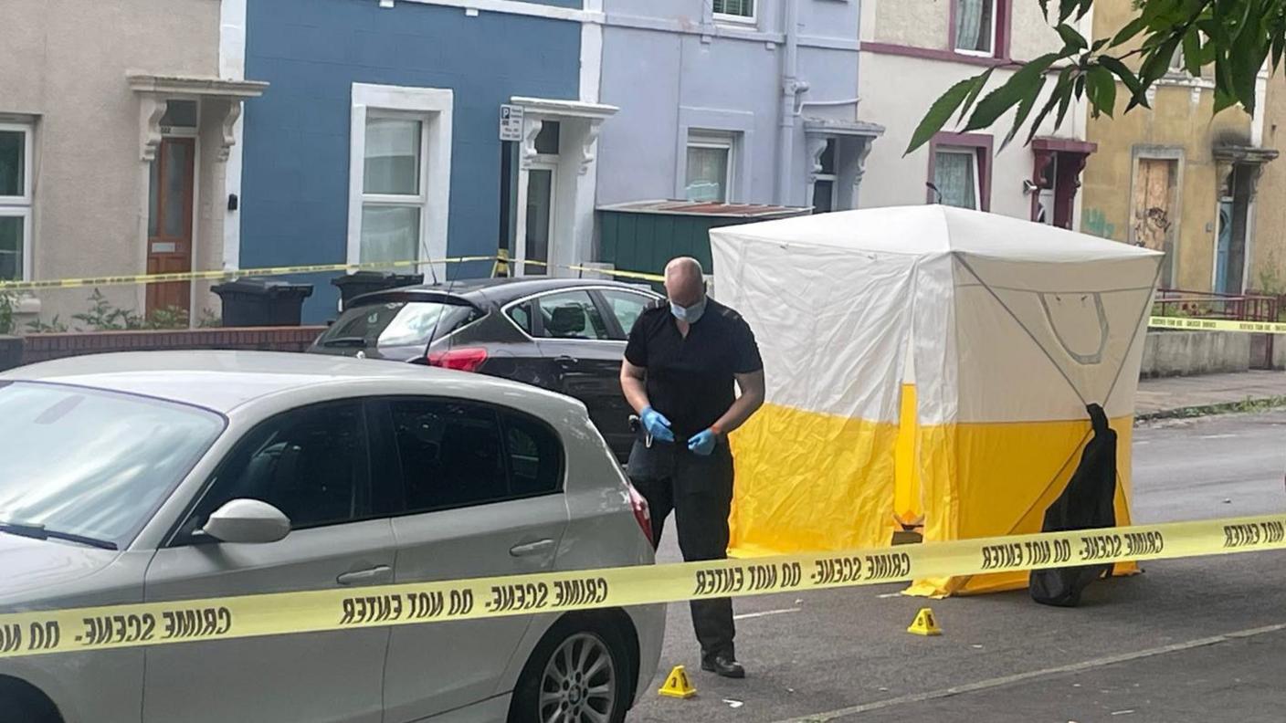 A police officer at the scene with yellow cones on the floor. He is standing with blue gloves and a face mask on. A yellow and white police tent is pictured behind him and there is a police cordon around the street.