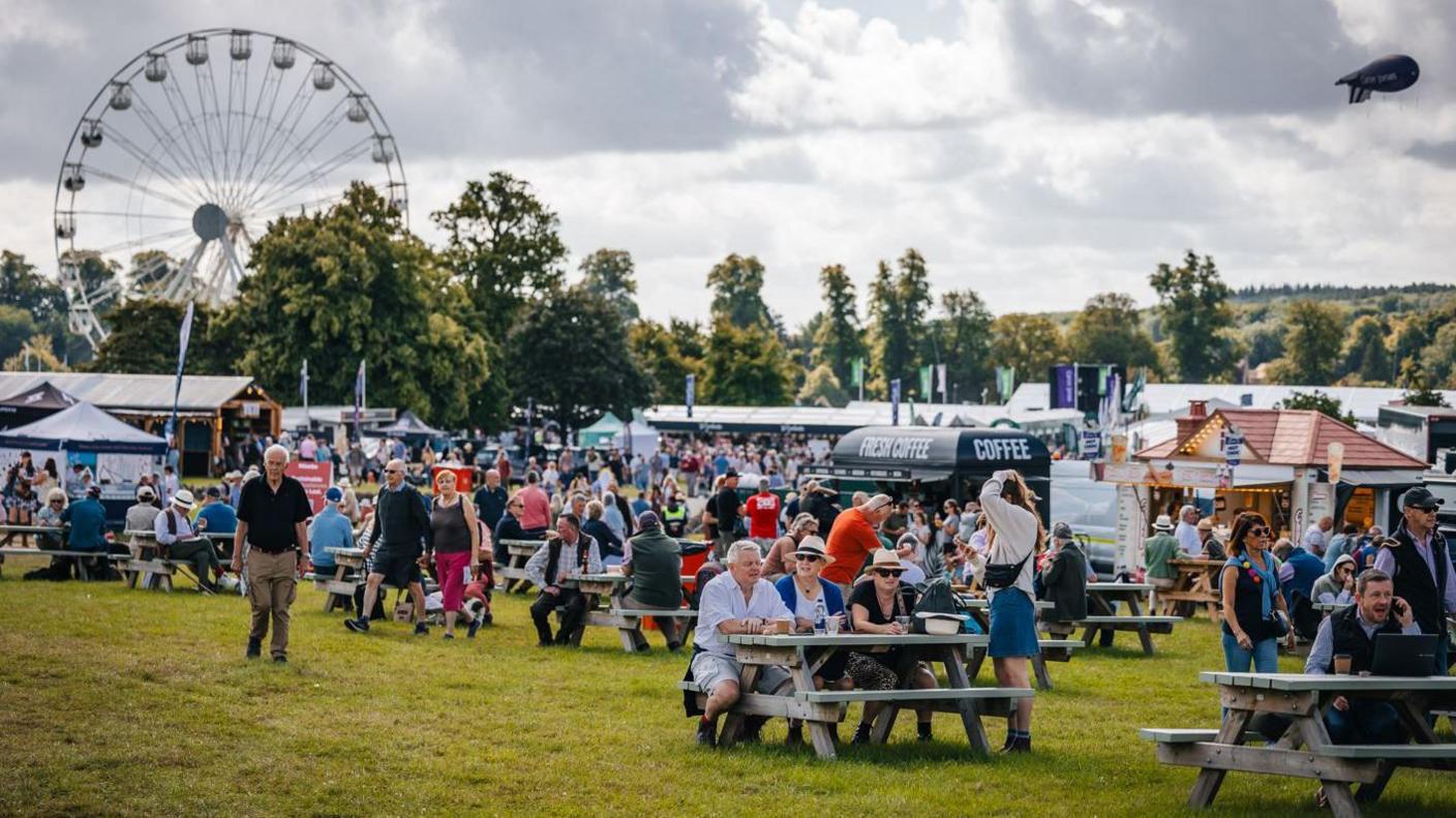 A large gathering of people at an open-air event, with some sitting on wooden picnic benches in the foreground and stalls and a big wheel in the background