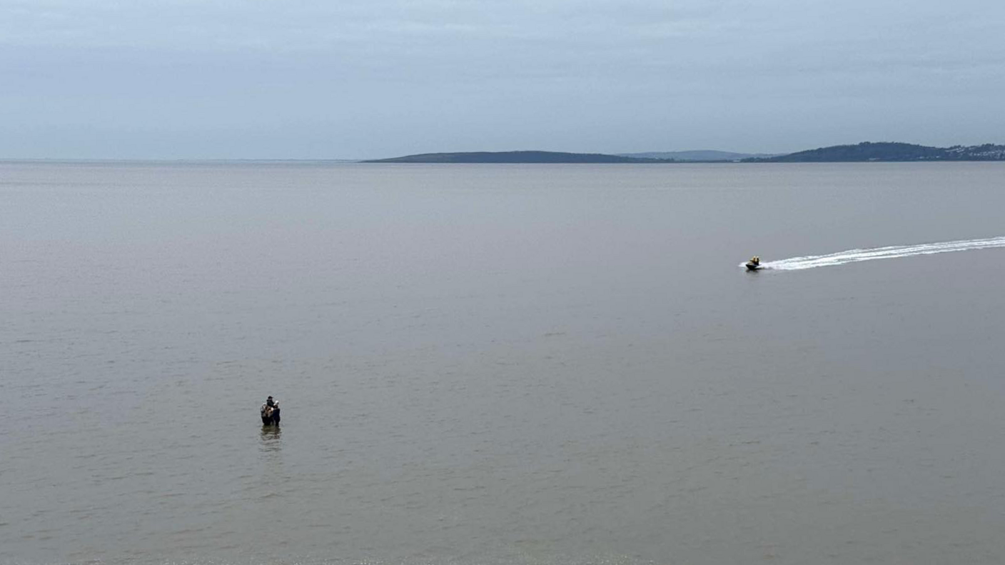 People stand huddled together, knee deep in water, as a speedboat races towards them.