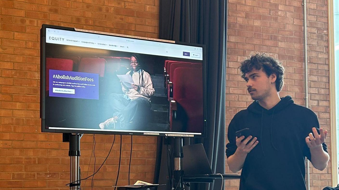 On the left of the image, there is a screen with a presentation on it. You can read  #AbolishAuditionFees in a purple box to the left of the screen and on the right sits a woman in a theatre. On the right of the image, Joshua Bendall, chair of Equity's student deputies committee, stands looking at the screen and presenting. He is wearing a black hoodie. 