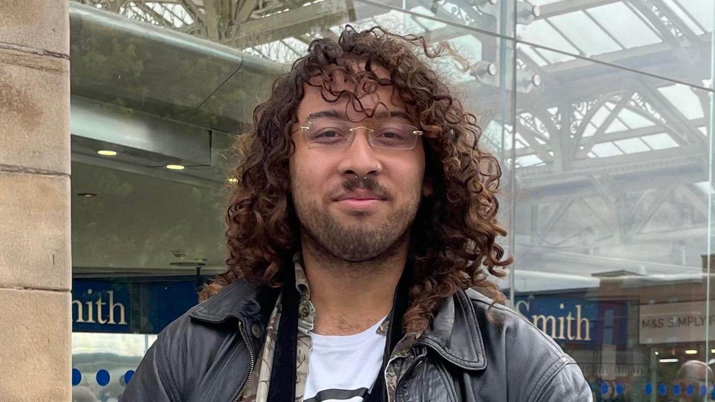 A man in his early 20s with curly dark brown hair, just past his shoulders. He wears glasses with no frame, two nose piercings, and has a short black beard. He is wearing a black leather jacket over a patterned shirt and white t-shirt.