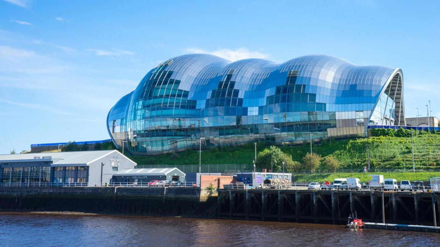 The Glasshouse in Gateshead. The bubble-like building is made of glass and overlooks the River Tyne. It sits on a hill covered in grass and plants.