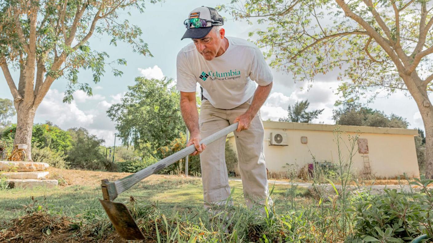 Simon King, in a white T-shirt, baseball hat and beige trousers, wields a gardening tool as he tidies the kibbutz gardens. He is surrounded by greenery.