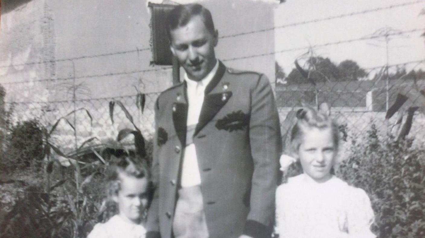 A black and white image of a man in a military-style jacket holding the hands of his two daughters in white blouses.