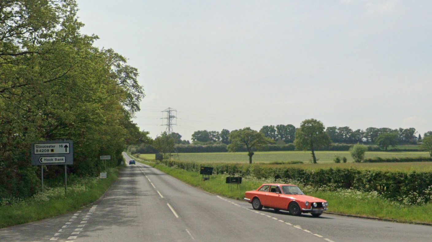 The B4208 road on a sunny day. The road is lined on one side with hedgerows and the other with trees. A car is visible in the distance, with another car in the foreground turning off the road at a junction.