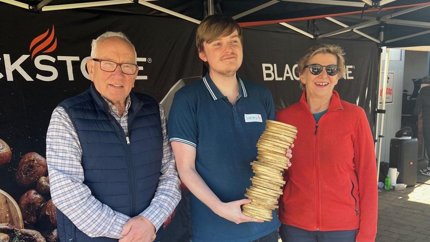 3 people are standing next to each other. The one on the left has a blue chequered shirt on, and a blue body warmer. He has glasses and silver hair. The one in the middle has brown hair, and is smiling towards the camera. He is holding the golden stack of pancakes. The woman on the right is wearing a red top, and has brown hair and sunglasses on. 