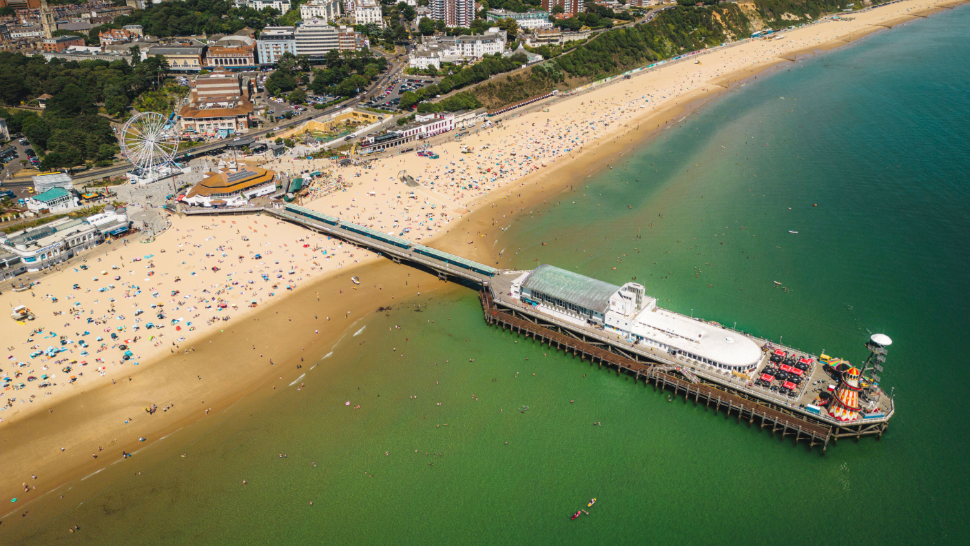 Aerial image of Bournemouth Pier with a large white building on it, a helter skelter and a high platform attached to a zip wire. The beach is busy and there is a big wheel on the pier approach.
