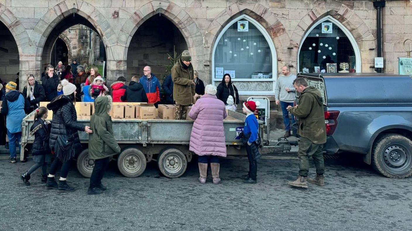 A farming trailer, carrying many food donation boxes, is pulled up outside a stone building. There are children and people looking in the trailer. 