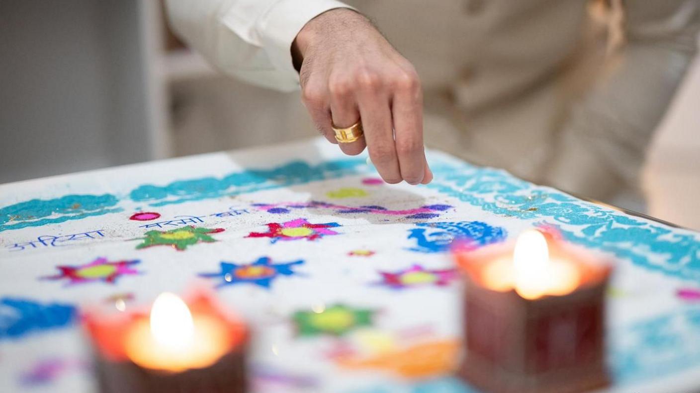 A plate with coloured powder in made in to flower designs. Dev is gently dropping the powder to create the detailing. Two candles are in front of it.