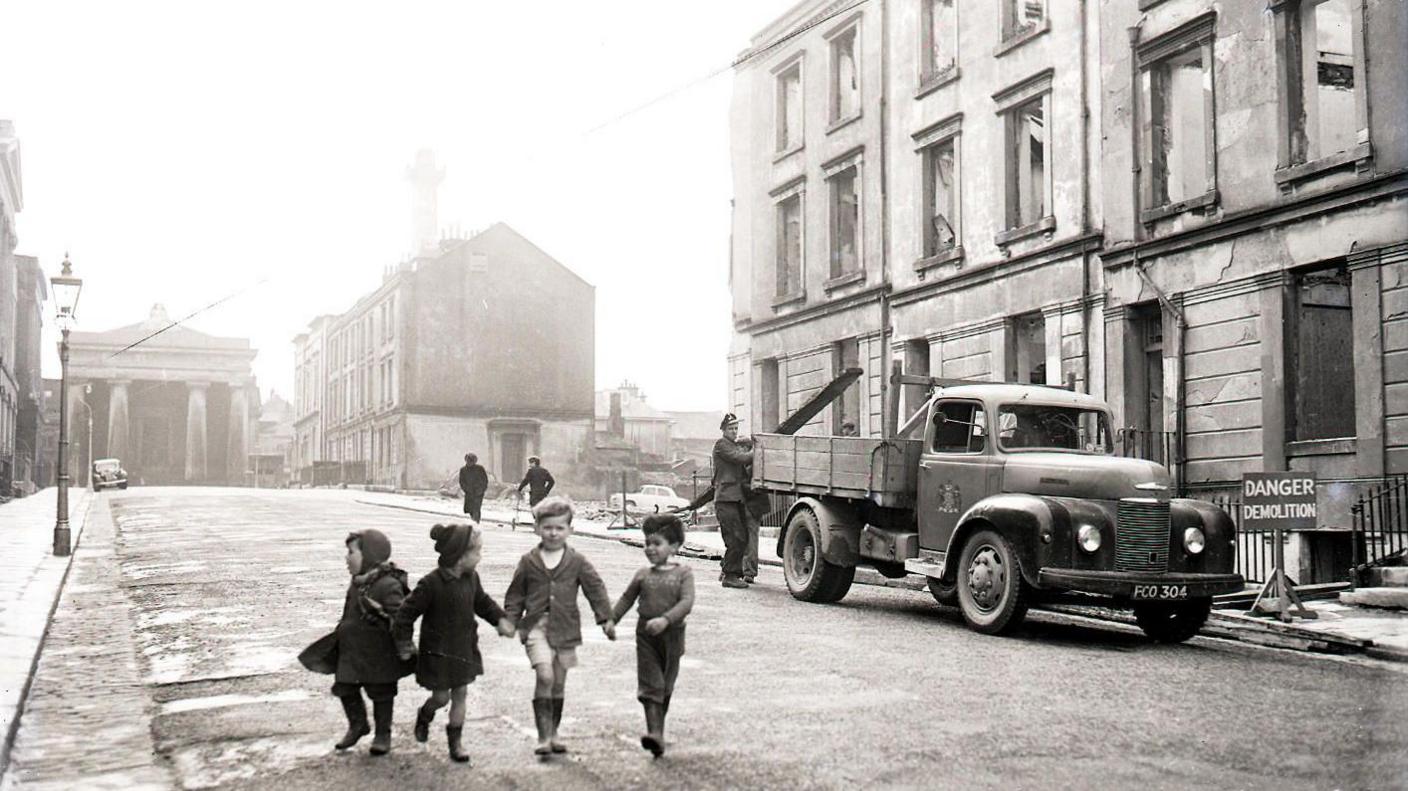 A black and white image shows four children running down Ker Street holding hands. There's a gap in the houses along the terraced street where buildings have been bombed. A truck is be loaded with wood from the remaining houses that are due to be demolished.