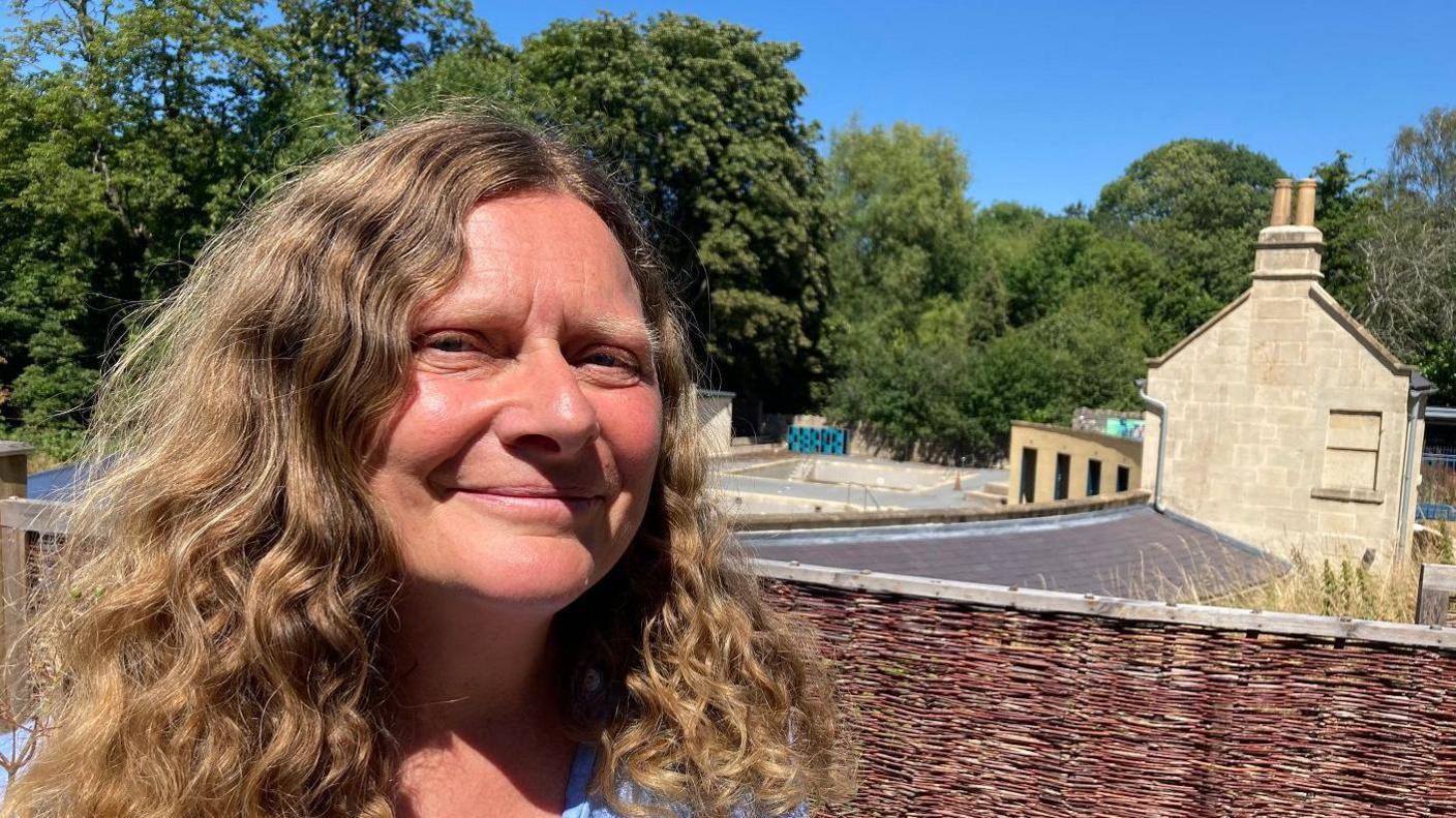 Close up of Karen's face, she has light brown, curly hair. She is wearing a blue shirt. The pool is in the background of the image.