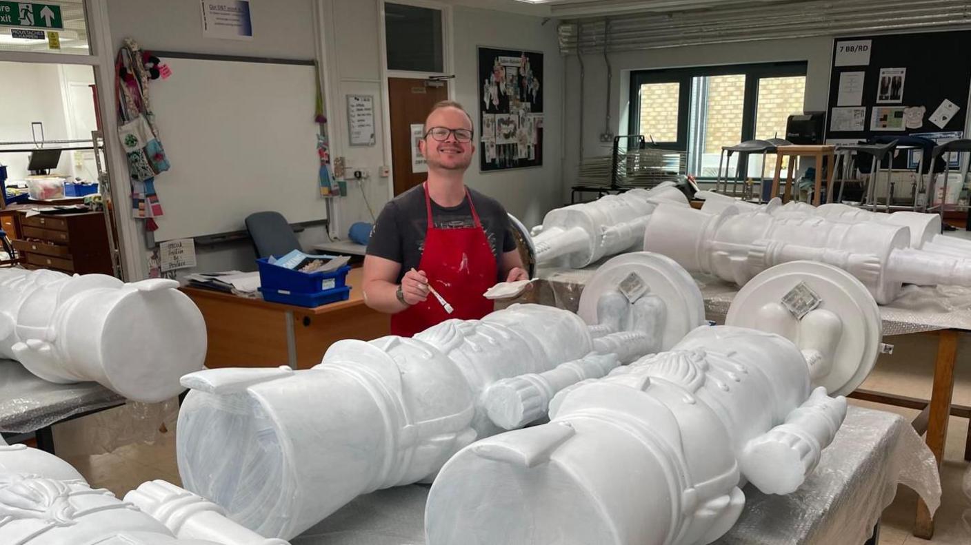 Marc Godfrey-Murphy in a red apron smiling while painting a number of Nutcracker sculptures as they lay down on tables. 