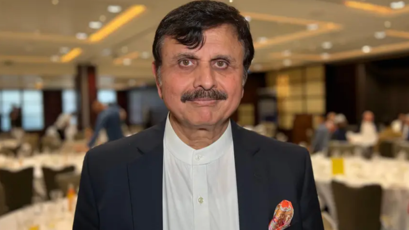 Dr Sarfaraz Jamali wears a white shirt and black blazer as he smiles toward the camera with an event including tables and guests taking place behind him