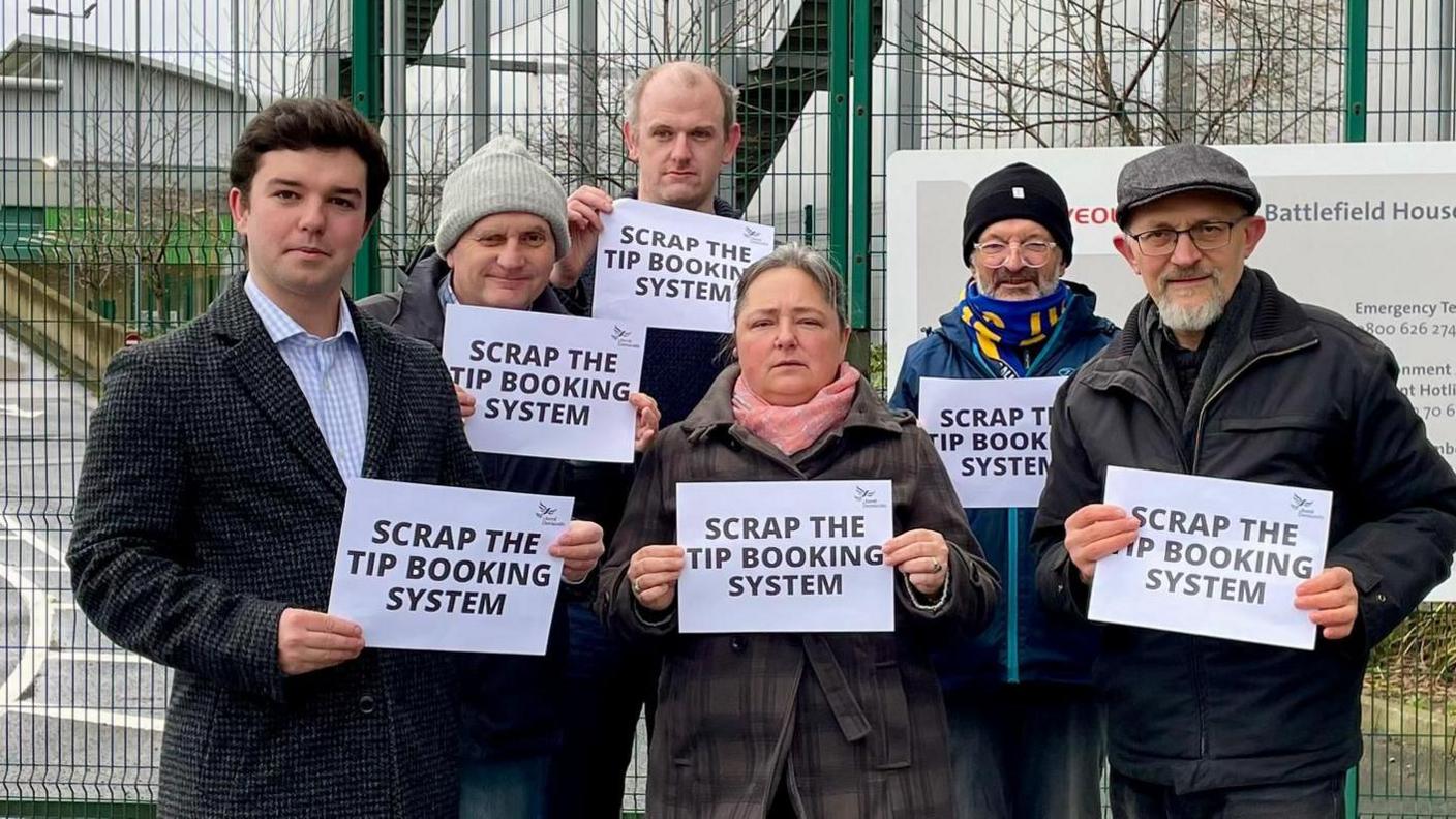 Five men and a woman wearing winter clothing posing in front of green gates, holding up white A4 pieces of paper with the words "scrap the tip booking system"